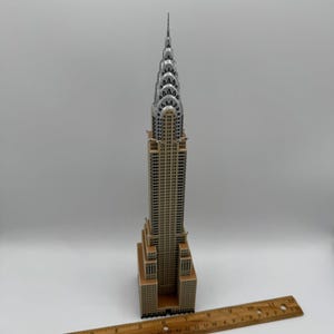 May include: A detailed model of the Chrysler Building, featuring a tan and silver color scheme. The model is placed on a white surface with a wooden ruler nearby. The building's iconic Art Deco design is clearly visible.