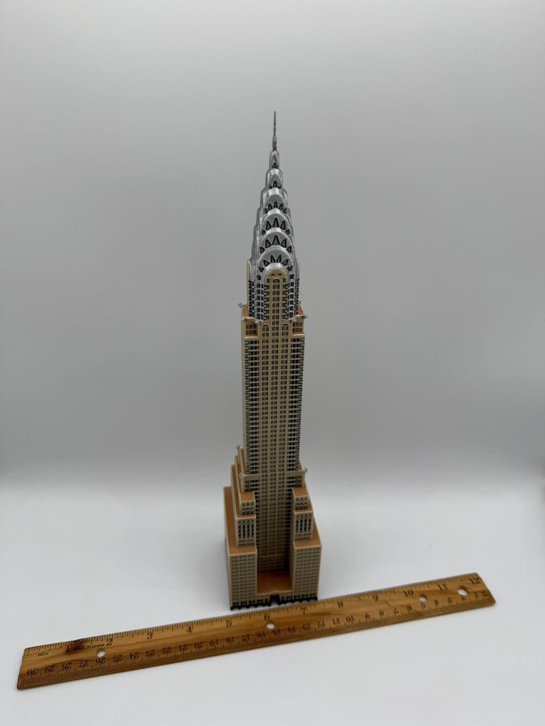 May include: A detailed model of the Chrysler Building, featuring a tan and silver color scheme. The model is placed on a white surface with a wooden ruler nearby. The building's iconic Art Deco design is clearly visible.