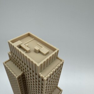 May include: A beige, 3D-printed model of a skyscraper. The building has multiple tiers and a flat roof with architectural details. The windows are small and evenly spaced. The model is set against a plain white background.