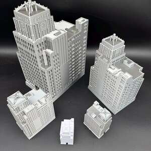 May include: A collection of gray miniature building models, each with intricate architectural details. The models vary in size and shape, showcasing different building designs. The word "EASTERN" is visible on one of the buildings. The buildings are arranged on a dark surface.