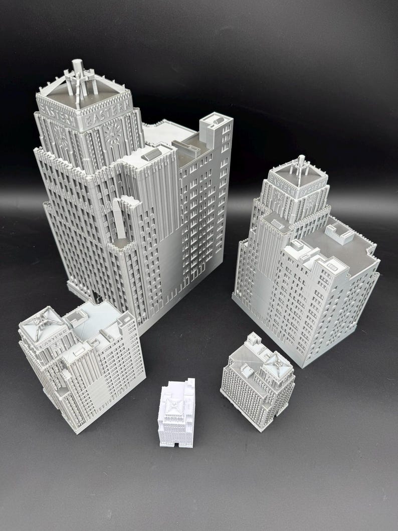 May include: A collection of gray miniature building models, each with intricate architectural details. The models vary in size and shape, showcasing different building designs. The word "EASTERN" is visible on one of the buildings. The buildings are arranged on a dark surface.