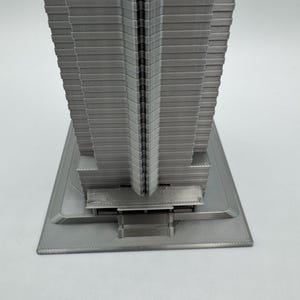 May include: A gray, 3D-printed model of a skyscraper. The building has a rectangular base and a tall, multi-tiered structure. The model is made of a solid, light gray material and has a detailed, textured surface.