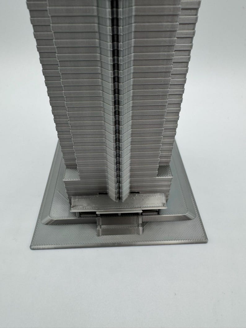 May include: A gray, 3D-printed model of a skyscraper. The building has a rectangular base and a tall, multi-tiered structure. The model is made of a solid, light gray material and has a detailed, textured surface.