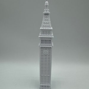 May include: A white 3D printed model of a tall building with a pointed roof and many windows. The building has a clock face on the front.