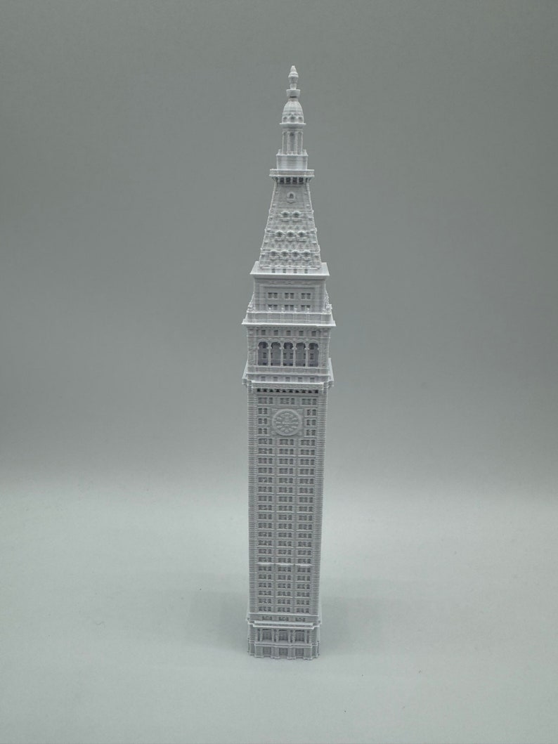 May include: A white 3D printed model of a tall building with a pointed roof and many windows. The building has a clock face on the front.