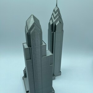 May include: Two gray 3D printed models of skyscrapers. The taller building has a pointed top and the shorter building has a flat top.