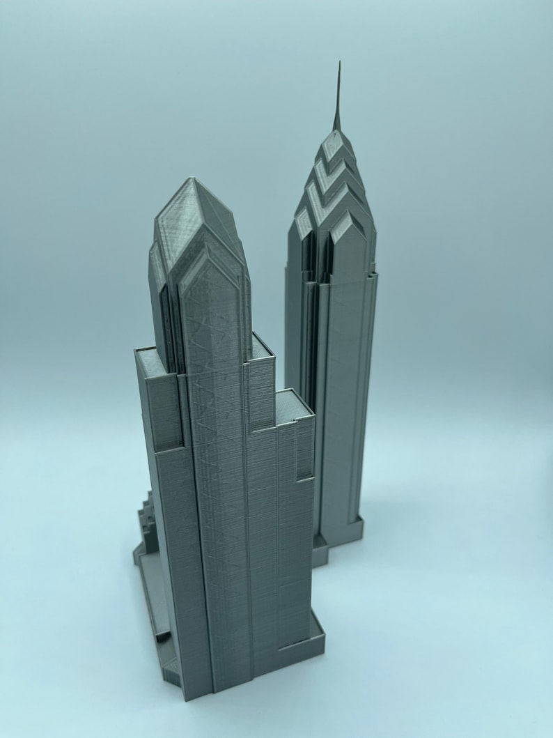 May include: Two gray 3D printed models of skyscrapers. The taller building has a pointed top and the shorter building has a flat top.