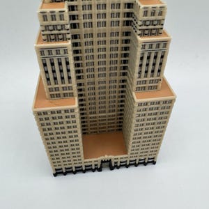 May include: A beige and brown 3D-printed model of a skyscraper. The building features numerous windows and architectural details, with a rectangular base and multiple tiers. The model is set against a white background.