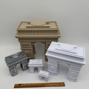 Może przedstawiać: Kolekcja miniaturowych modeli Łuku Triumfalnego w r&oacute;żnych rozmiarach i kolorach. Modele są w kolorze beżowym, szarym i białym. Drewniana linijka umieszczona przed modelami. Są to repliki architektoniczne.