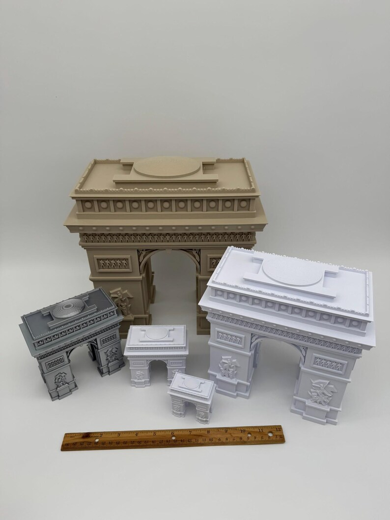 Może przedstawiać: Kolekcja miniaturowych modeli Łuku Triumfalnego w r&oacute;żnych rozmiarach i kolorach. Modele są w kolorze beżowym, szarym i białym. Drewniana linijka umieszczona przed modelami. Są to repliki architektoniczne.