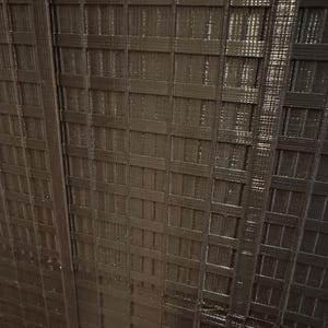 May include: Close-up of a dark brown, textured surface with a repeating grid pattern. The pattern consists of rectangular and square shapes, creating a woven or lattice-like appearance. The surface has a slightly glossy finish.