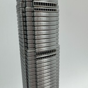 May include: A detailed, silver-colored model of a skyscraper. The building features a ribbed facade with horizontal bands and rectangular vent-like details. The model is a vertical structure, showcasing architectural design.