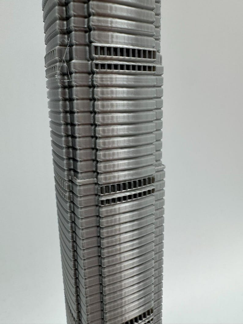 May include: A detailed, silver-colored model of a skyscraper. The building features a ribbed facade with horizontal bands and rectangular vent-like details. The model is a vertical structure, showcasing architectural design.