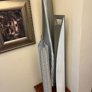 May include: Three silver 3D printed models of skyscrapers on a wooden table. The tallest model is a curved building, the middle model is a tall, thin building with a pointed top, and the shortest model is a rectangular building with a large opening in the middle.