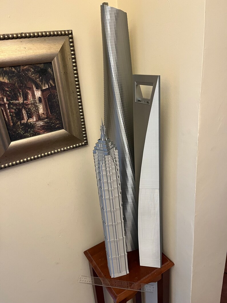 May include: Three silver 3D printed models of skyscrapers on a wooden table. The tallest model is a curved building, the middle model is a tall, thin building with a pointed top, and the shortest model is a rectangular building with a large opening in the middle.