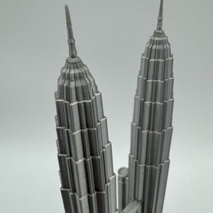 May include: A 3D printed model of the Petronas Towers in Kuala Lumpur, Malaysia. The model is made of a silver-colored plastic and is detailed with the towers' distinctive spires and windows.