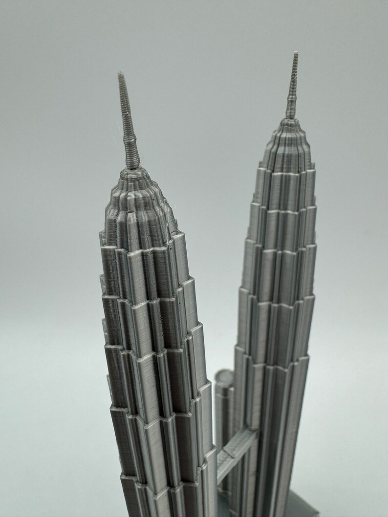 May include: A 3D printed model of the Petronas Towers in Kuala Lumpur, Malaysia. The model is made of a silver-colored plastic and is detailed with the towers' distinctive spires and windows.