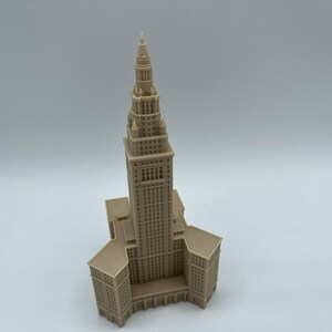May include: A beige 3D printed model of a tall building with a pointed spire. The building has a rectangular base and a series of windows and details.