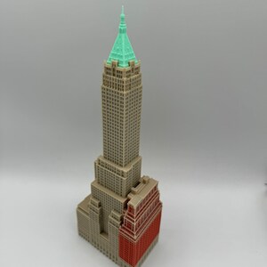 May include: A miniature model of a skyscraper with a green roof and a red brick base. The building is made of beige plastic and has many windows.