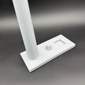 May include: A light gray 3D-printed stand with a rectangular base, a cylindrical support, a small cone, and a square cutout. The stand is designed to hold an item, likely a phone or small device.