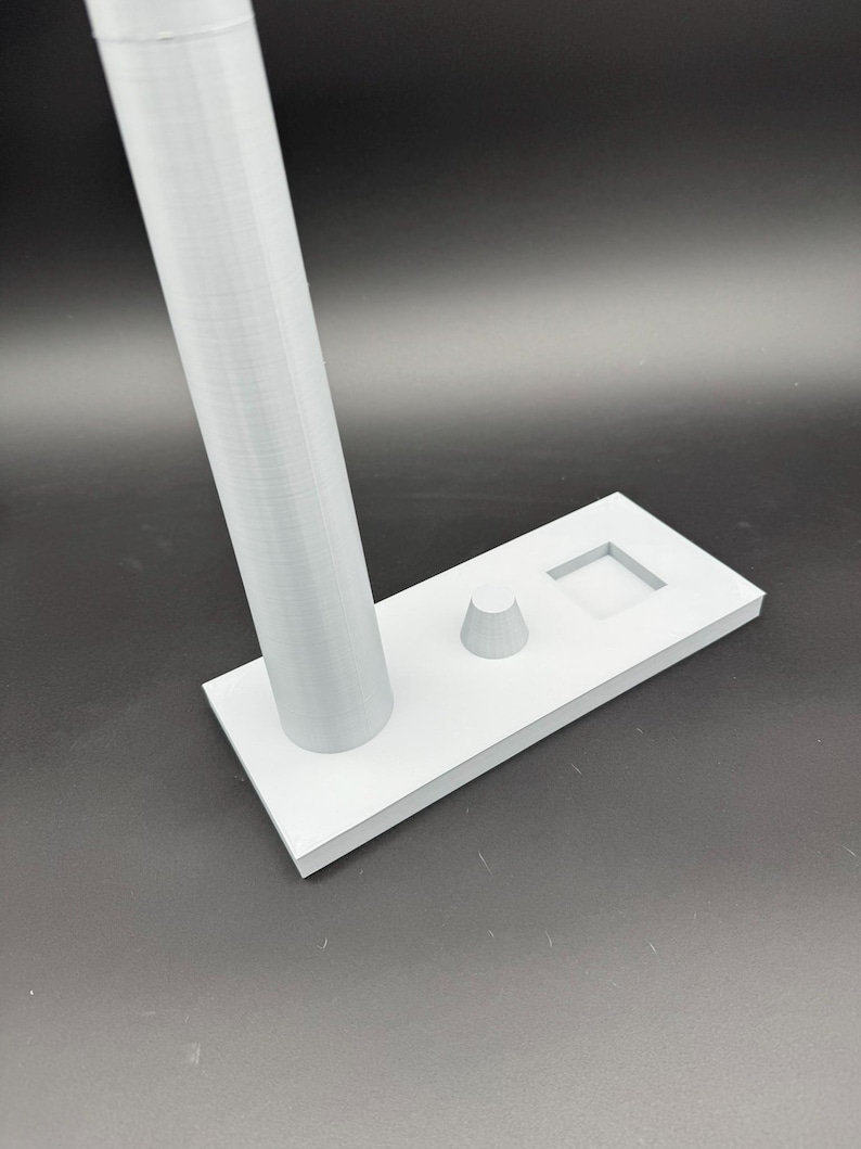 May include: A light gray 3D-printed stand with a rectangular base, a cylindrical support, a small cone, and a square cutout. The stand is designed to hold an item, likely a phone or small device.