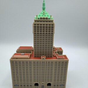 May include: A detailed model of a skyscraper with a tan facade, red rooftops, and a green-topped tower. The building features numerous windows and architectural details, showcasing a classic design.