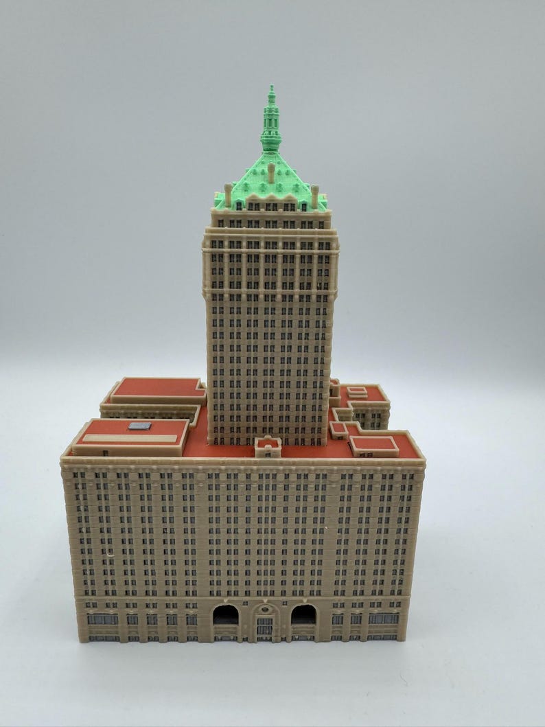 May include: A detailed model of a skyscraper with a tan facade, red rooftops, and a green-topped tower. The building features numerous windows and architectural details, showcasing a classic design.