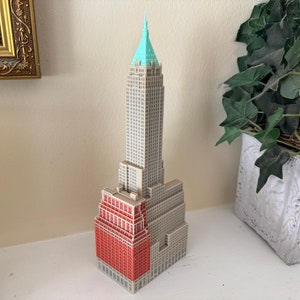 May include: A miniature model of a tall, grey building with a red brick facade and a green roof. The building is on a white surface.