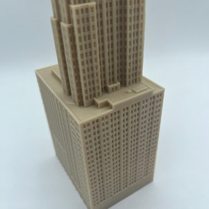 May include: A detailed model of a skyscraper, made of a light brown plastic material. The model is a miniature replica of a tall building with many windows and a flat roof.