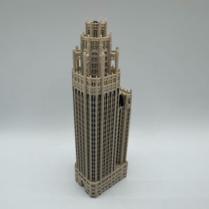 May include: A beige, three-dimensional architectural model of a skyscraper featuring a stepped, multi-tiered design with intricate vertical detailing, arched window patterns, and a central tower structure that tapers toward the top.