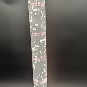 May include: A tall, gray skyscraper lamp with a pixelated design. The lamp features white and pink illuminated windows, creating a modern and architectural aesthetic. The lamp is plugged in and has a white cord.