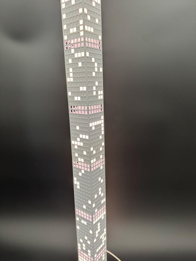 May include: A tall, gray skyscraper lamp with a pixelated design. The lamp features white and pink illuminated windows, creating a modern and architectural aesthetic. The lamp is plugged in and has a white cord.