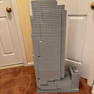 May include: A gray model of a skyscraper with a multi-tiered design. The building has many small, square windows and a rectangular base. The model is made of a light gray material and stands tall against a neutral background.