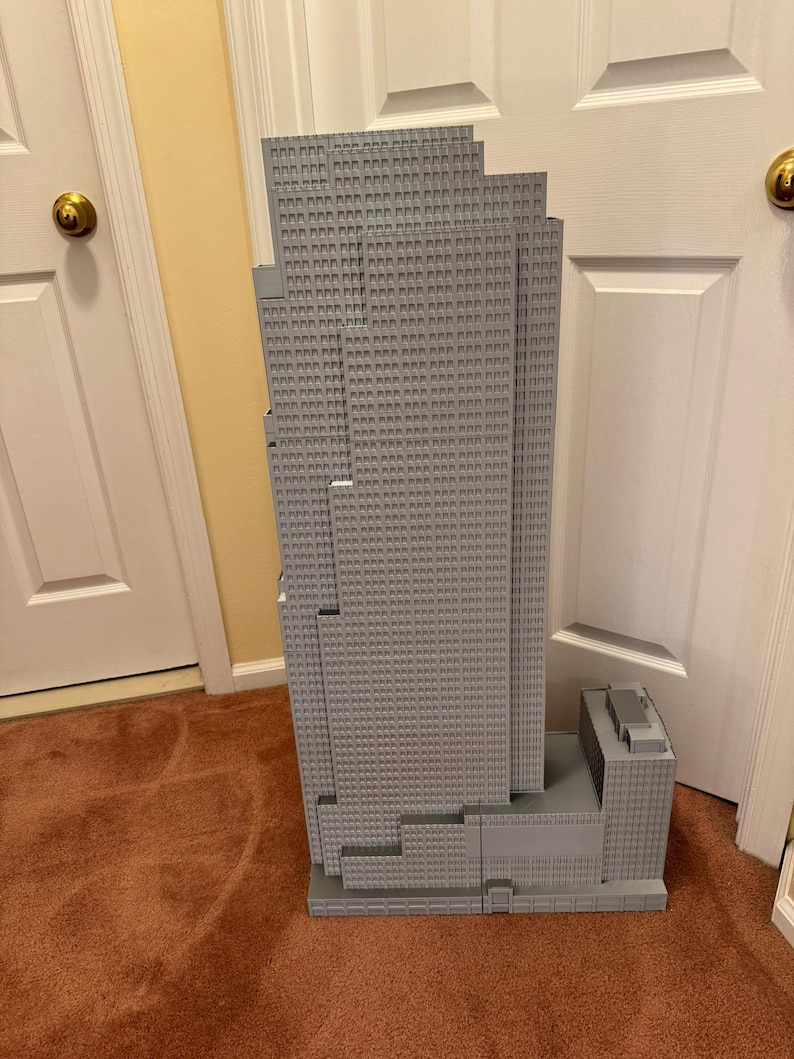 May include: A gray model of a skyscraper with a multi-tiered design. The building has many small, square windows and a rectangular base. The model is made of a light gray material and stands tall against a neutral background.