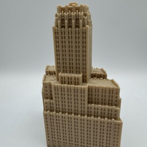 May include: A tan-colored miniature skyscraper model with a detailed facade. The building has multiple tiers and a tall central tower, featuring numerous windows and architectural details. The model is likely made of plastic or resin.
