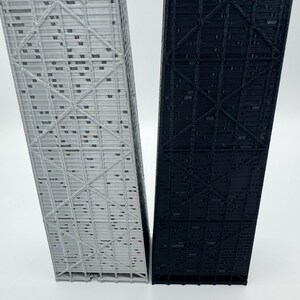 May include: Two architectural models, one light gray and one black, stand side-by-side. The models are rectangular with intricate, repeating geometric patterns. The structures appear to be miniature representations of skyscrapers or similar buildings.