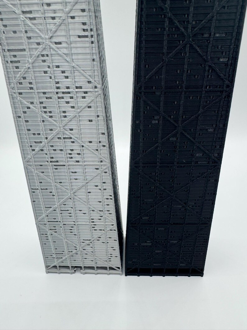 May include: Two architectural models, one light gray and one black, stand side-by-side. The models are rectangular with intricate, repeating geometric patterns. The structures appear to be miniature representations of skyscrapers or similar buildings.