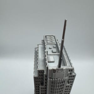 May include: A gray, 3D-printed model of a skyscraper. The building has a detailed facade with numerous windows and architectural features. A tall antenna extends from the top of the building. The model is set against a plain, light gray background.
