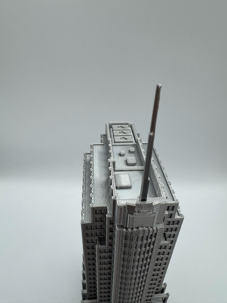 May include: A gray, 3D-printed model of a skyscraper. The building has a detailed facade with numerous windows and architectural features. A tall antenna extends from the top of the building. The model is set against a plain, light gray background.