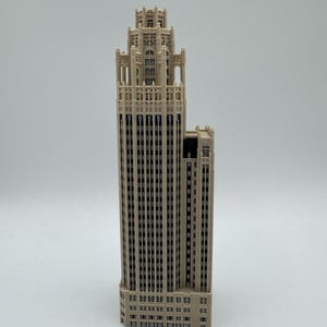 May include: A beige architectural model of a skyscraper featuring a tall, multi-tiered tower with intricate vertical detailing, numerous recessed windows, and a decorative crown at the top, standing upright against a plain light background.