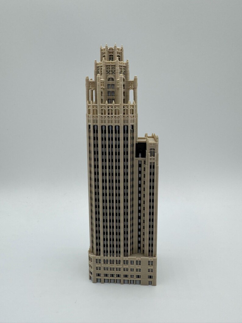 May include: A beige architectural model of a skyscraper featuring a tall, multi-tiered tower with intricate vertical detailing, numerous recessed windows, and a decorative crown at the top, standing upright against a plain light background.