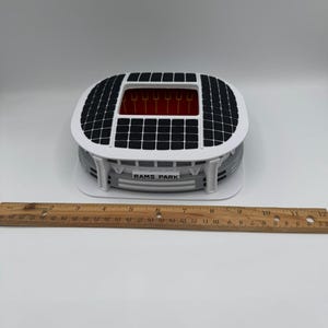 May include: A miniature stadium model with a white and gray exterior, a black roof, and a red interior. The model has the text "RAMS PARK" on the front. A wooden ruler is placed next to the model.
