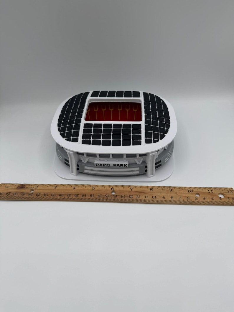May include: A miniature stadium model with a white and gray exterior, a black roof, and a red interior. The model has the text "RAMS PARK" on the front. A wooden ruler is placed next to the model.