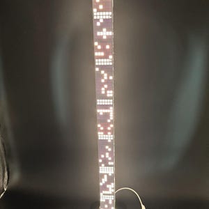 May include: A tall, rectangular LED lamp with a black base. The lamp's body is a series of illuminated white squares against a purple background, resembling a skyscraper. A white cord extends from the base.