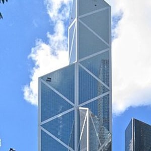 May include: A tall, modern skyscraper with a unique geometric design. The building features a white frame with triangular and rectangular glass panels. The sky is blue with fluffy white clouds, and green trees are visible at the base of the building.