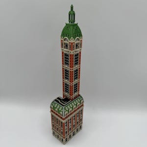 May include: A miniature model of a skyscraper featuring a tall, narrow tower with a green dome roof. The structure has red brick-colored walls, white architectural detailing, and multiple rows of black windows, set on a wider rectangular base.