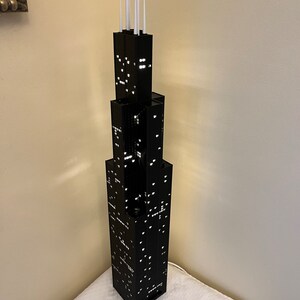 May include: A black LEGO model of the Willis Tower, formerly known as the Sears Tower, with white antenna details. The model is lit up with white lights, creating a realistic city skyline effect.