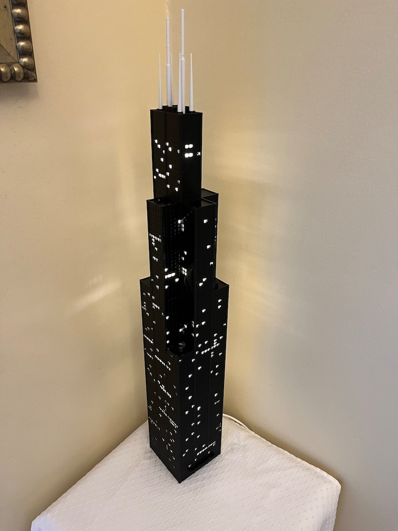 May include: A black LEGO model of the Willis Tower, formerly known as the Sears Tower, with white antenna details. The model is lit up with white lights, creating a realistic city skyline effect.