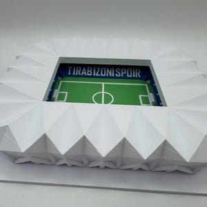 May include: A white, geometric model of a stadium with a green field and the text "TIRABZONISPOIR" visible. The stadium has a detailed, angular design, and the field features white markings.