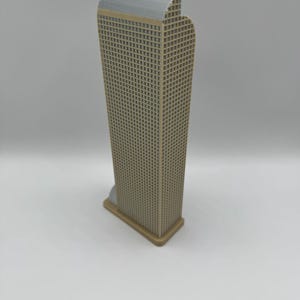 May include: A beige and gray miniature skyscraper model. The building has a rectangular shape with a grid pattern representing windows. The top of the building is gray, and the base is a lighter beige color. The model is standing on a white surface.